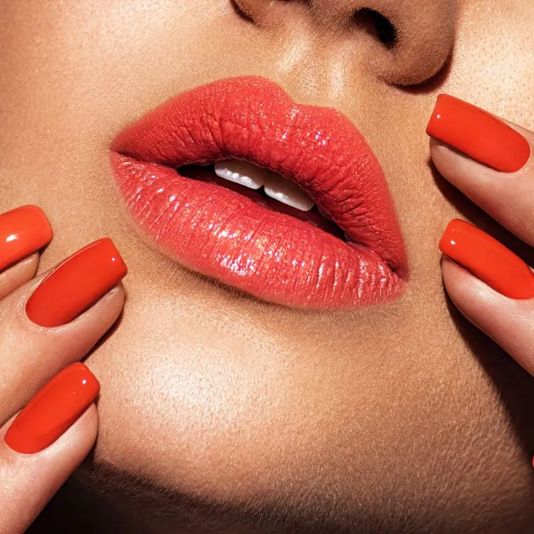 A imagem mostra um close-up dos lábios de uma pessoa, pintados de vermelho brilhante. As unhas também estão visíveis, pintadas com esmalte vermelho combinando. A pele parece lisa e a imagem tem um tom quente.

