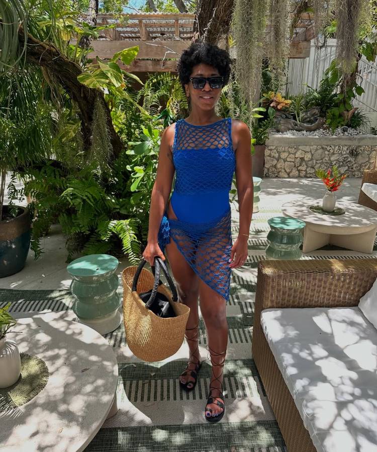 Uma mulher de óculos escuros, com cabelos curtos e cacheados, posa em uma área de descanso ao ar livre. Ela usa um maiô azul e uma saída de praia de crochê azul, com uma bolsa de palha grande e sandálias de amarrar.