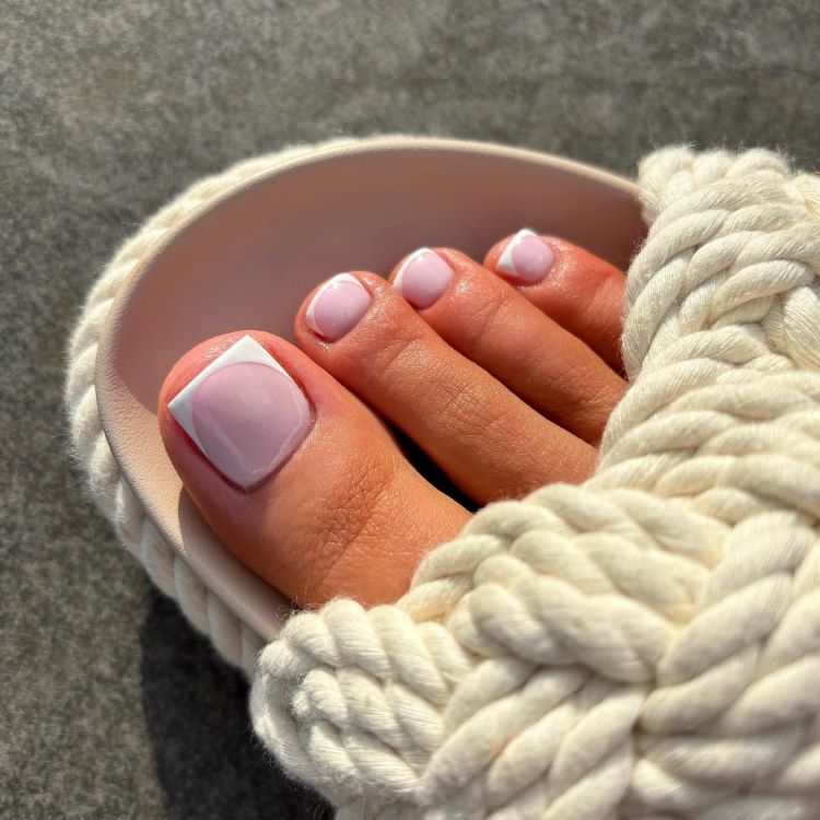 Pés femininos com unhas pintadas em esmalte azul claro e decoração de pequenas flores brancas. Sandálias de tiras finas em tom nude. Tornozeleira dourada delicada. Fundo de piso de madeira clara iluminado pelo sol, criando um visual elegante e descontraído.





