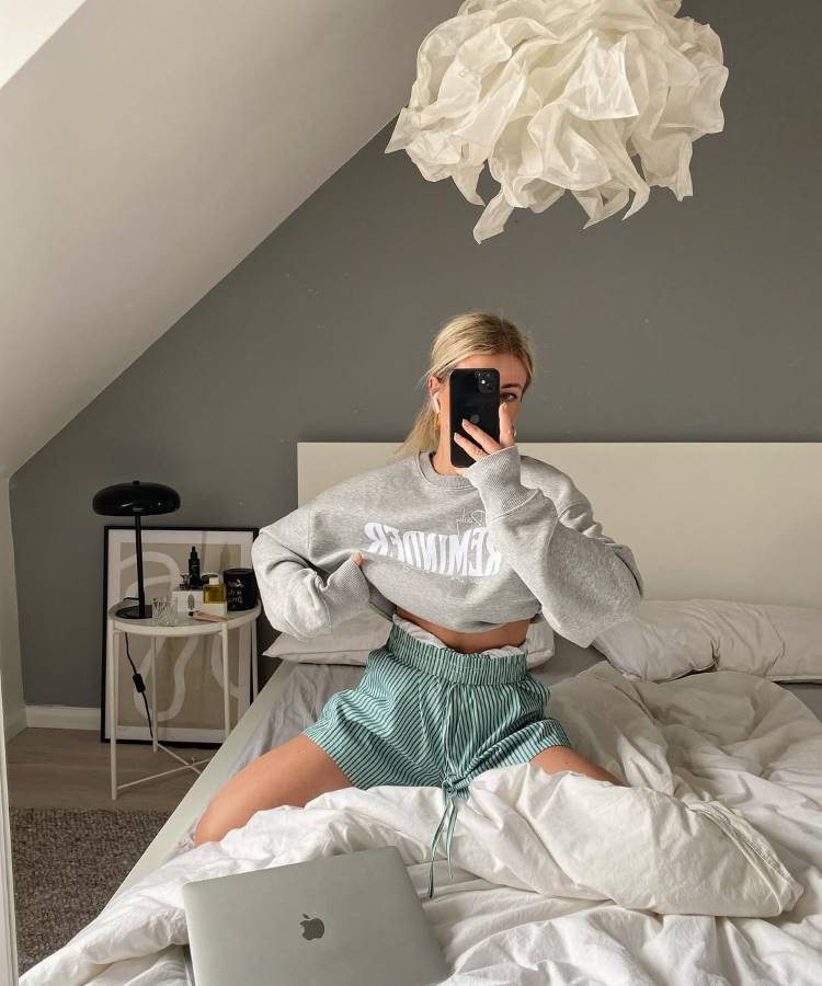Uma jovem loira de rabo de cavalo, vestindo um moletom cinza e shorts listrados. Ela se senta na cama com um cobertor branco e tira uma selfie em seu quarto com paredes cinzas e uma luminária de papel.
