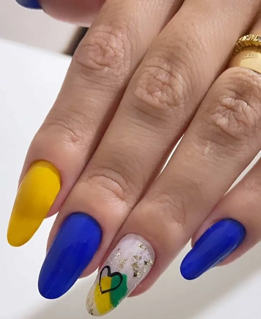 Imagem de mão com unhas decoradas para a Copa do Mundo 2022. Cores usadas, verde, amarelo, azul, branco e preto. Polegar, médio e mínimo, azul. Indicador, amarelo. Anelar, branco com verde e amarelo + desenho de coração preto + brilho dourado. 