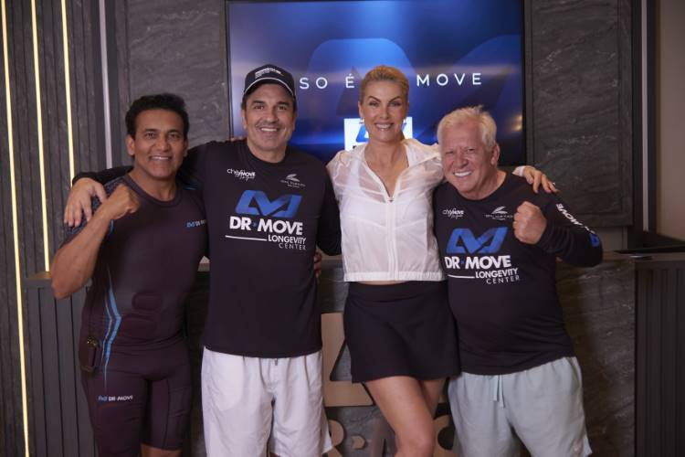 Quatro pessoas, duas mulheres e dois homens, sorriem para a câmera. Todos estão vestidos com roupas de treino da DR MOVE. A mulher loira usa shorts e um casaco branco. Eles estão na frente de uma tela grande com a marca da empresa.