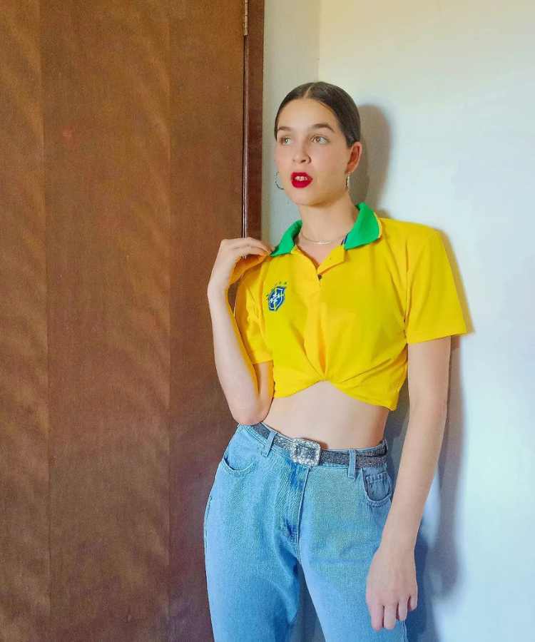 Imagem com fundo de porta e parede. No centro, mulher usando camiseta do Brasil + calça jeans com cinto