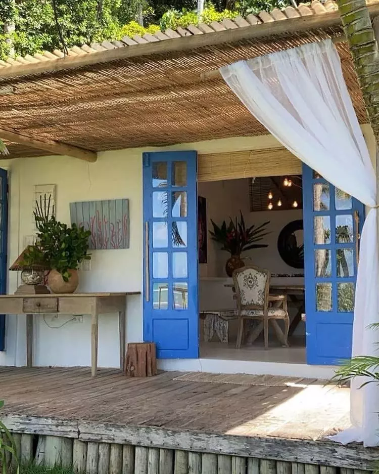 Frente de lar rural com porta azul. Alpendre com teto de palha com mesa de madeira e planta e luminária sob ela.