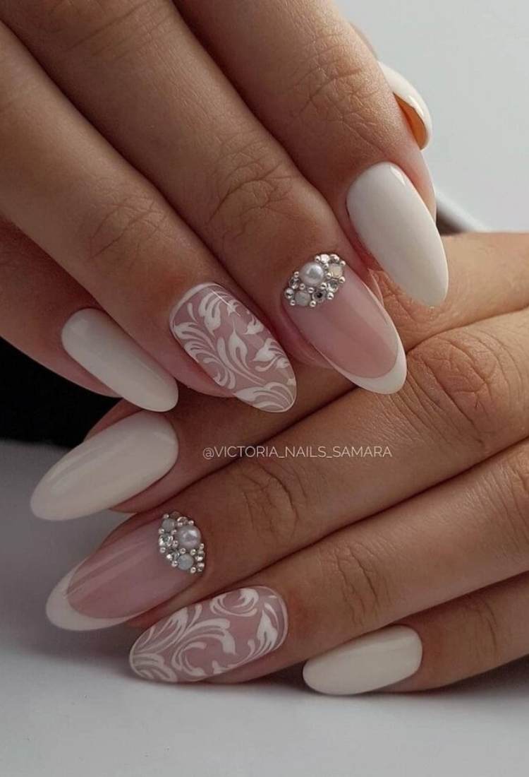 Unhas almond com indicador e mindinho brancas, médio com francesinha e pedraria e anelar com desenho de flores