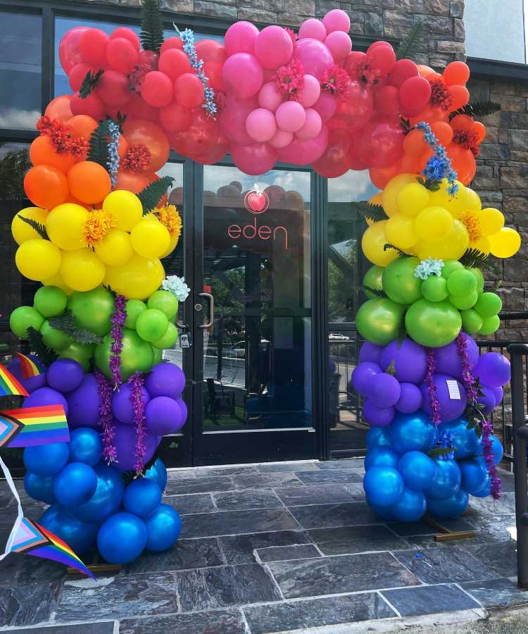 Arco decorativo feito de balões coloridos em formato de arco-íris com detalhes florais, em frente a uma entrada de vidro com a logo "eden". Bandeiras LGBTQIA+ complementam a cena, criando uma atmosfera vibrante e inclusiva.