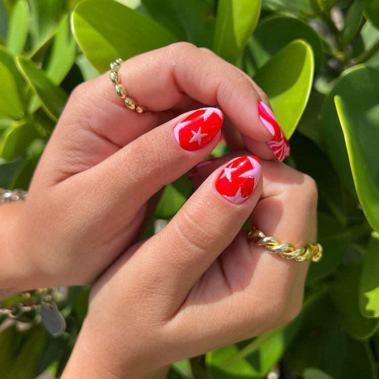 Mãos com estilos de unhas curtas, decoradas com esmalte vermelho vibrante e estrelas brancas, combinadas com detalhes em rosa ondulado. Dedos adornados com anéis dourados brilhantes. Fundo com folhas verdes iluminadas, criando um contraste tropical e moderno.