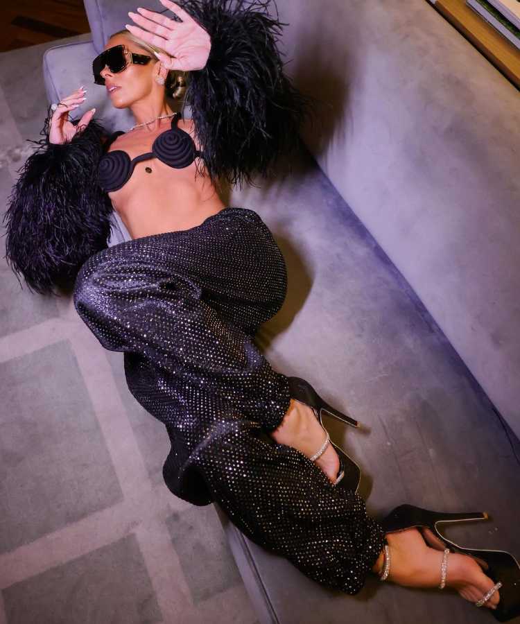 Looks de Adriane Galisteu no Carnaval 2023. Apresentadora no Baile da Narcisa usando top, luva de plumas e calça de paetês, pretos.