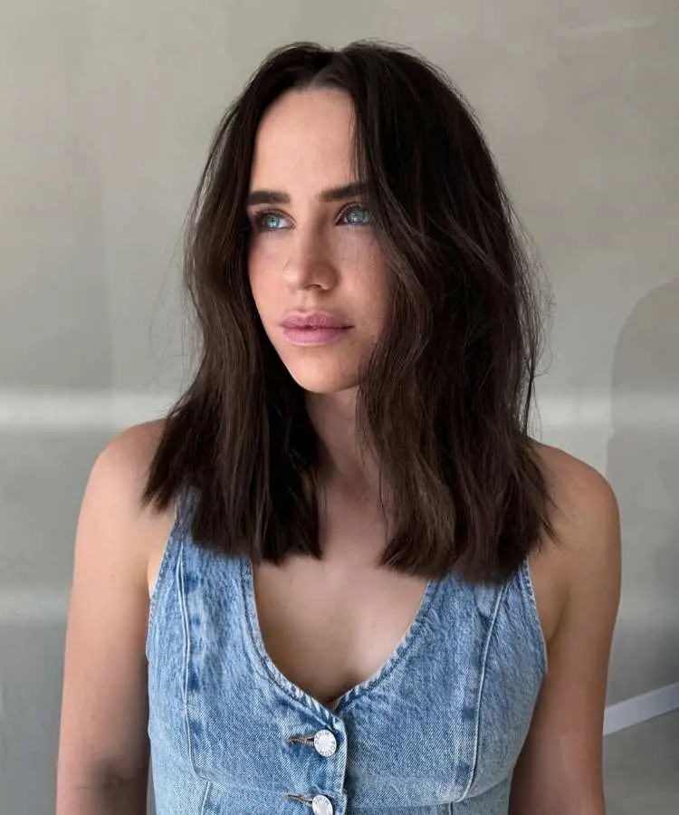 Mulher com cabelo médio castanho escuro, corte reto levemente ondulado, repartido ao meio, com textura natural. Ela usa um vestido jeans com botões e olha para o lado. Fundo claro e minimalista, destacando o visual clean e moderno.