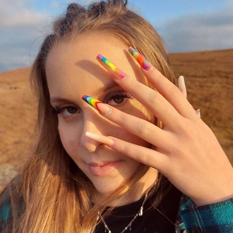 Mulher com a mão no rosto e unhas decoradas de Arco-íris: polegar: nude. Indicador: colorido mesclado. Médio: colorido com fio dourado. Anelar: arco-íris e pedrinhas na cutícula. Mínimo: nude.