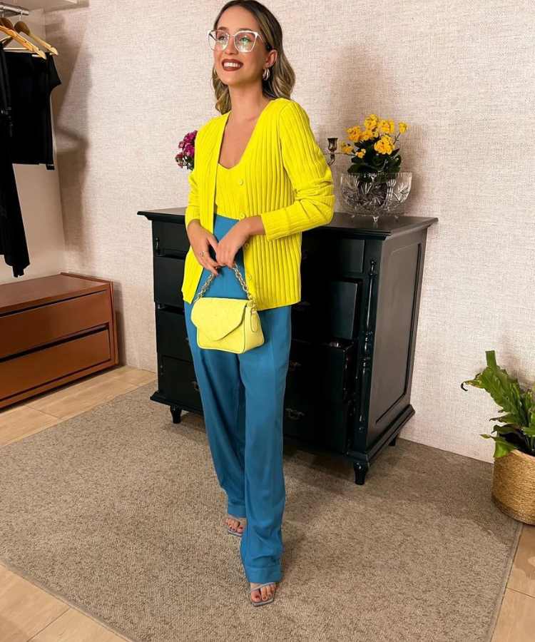 IMagem com fundo de parede e móvel preto. No centro, mulher usando suéter amarelo, calça azul, bolsa amarela