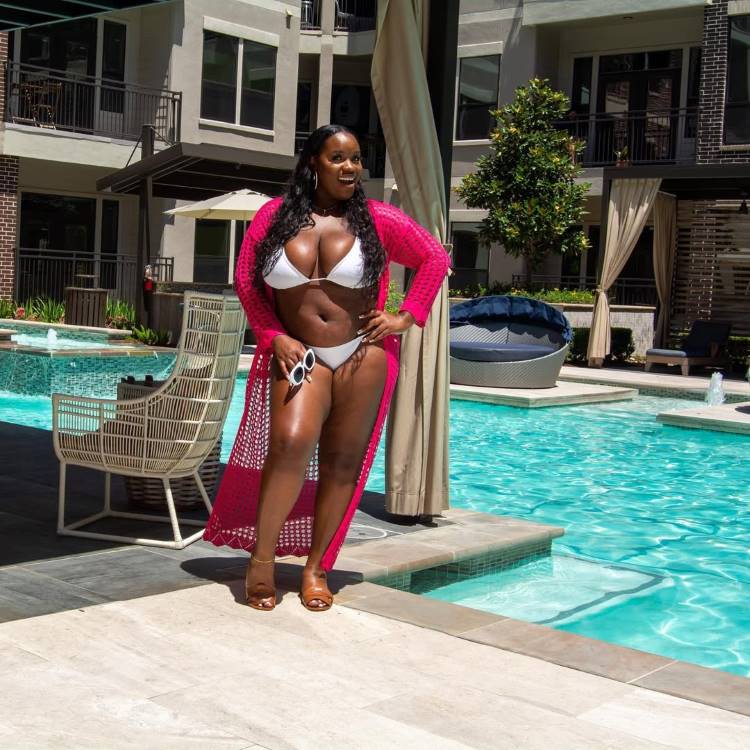 Mulher sorrindo em um deck de piscina, usando biquíni branco e uma longa saída de praia de crochê rosa. Ela posa com uma mão na cintura, segurando óculos de sol. Ao fundo, o prédio e a piscina.