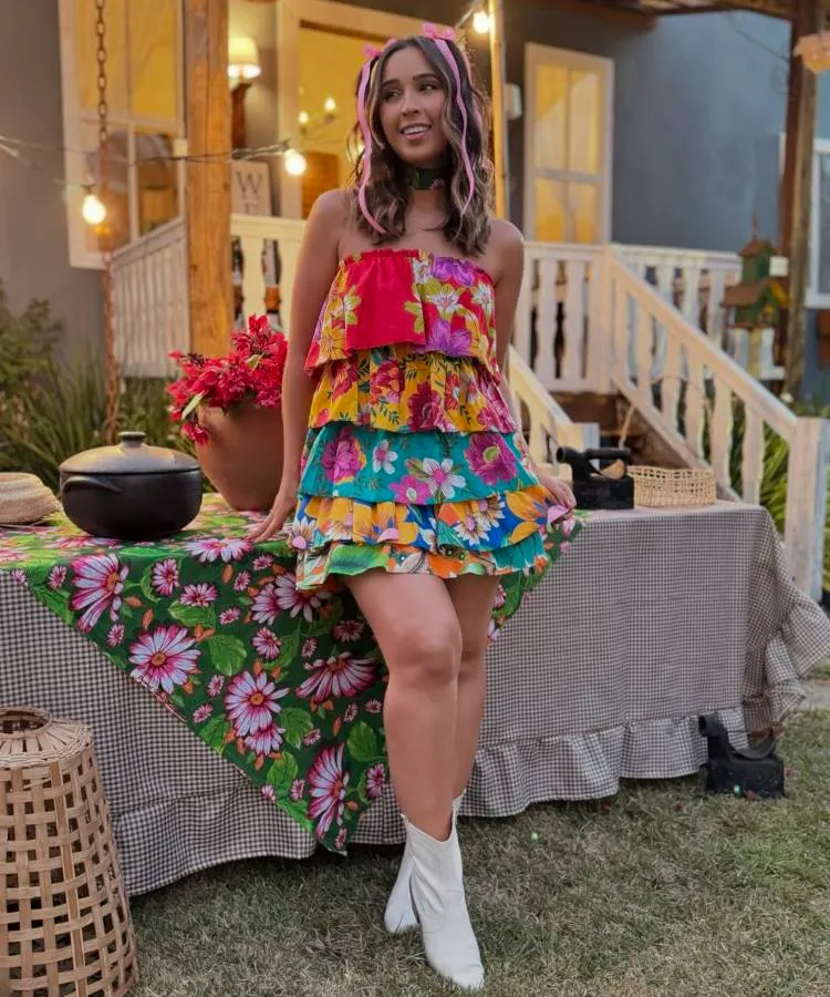 A imagem mostra uma mulher em ambiente minimalista, com cabelo preso por laço colorido combinando com sua roupa. Ela veste top peplum xadrez e jeans claros, com botas brancas. Segura bolsa vermelha, ao lado de espelho dourado e cadeira branca.
