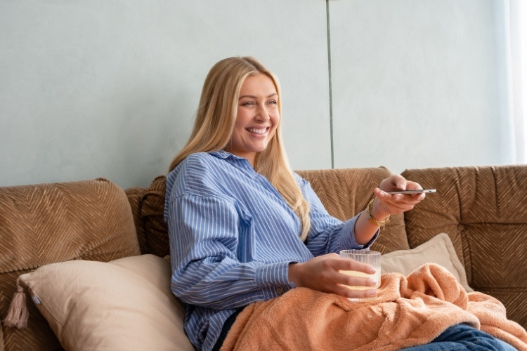 Mulher de pele clara de roupa azul no sofá tomando suco e mexendo no controle da TV 