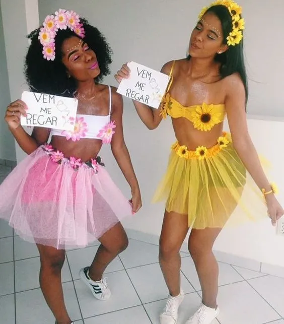 Duas mulheres de pele negra com top e saia de tule, uma rosa e outra amarela, com flores e placa "vem me regar"