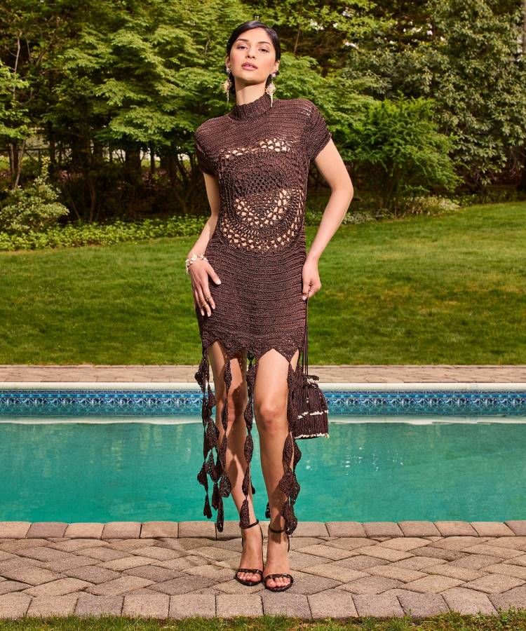 Uma mulher posa ao ar livre, perto de uma piscina, usando um vestido de crochê marrom escuro com detalhes vazados e franjas longas. Seus acessórios incluem brincos grandes e uma pequena bolsa combinando, realçando um visual boêmio e elegante.