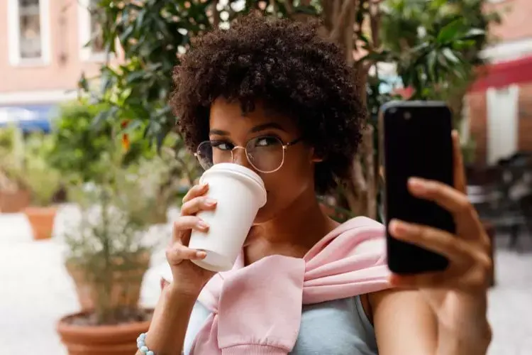 A imagem mostra uma mulher de cabelo cacheado, usando óculos, tirando uma selfie enquanto segura um copo de café. Ela veste uma blusa rosa sobre os ombros. Ao fundo, há plantas e um ambiente externo descontraído, com mesas e cadeiras.