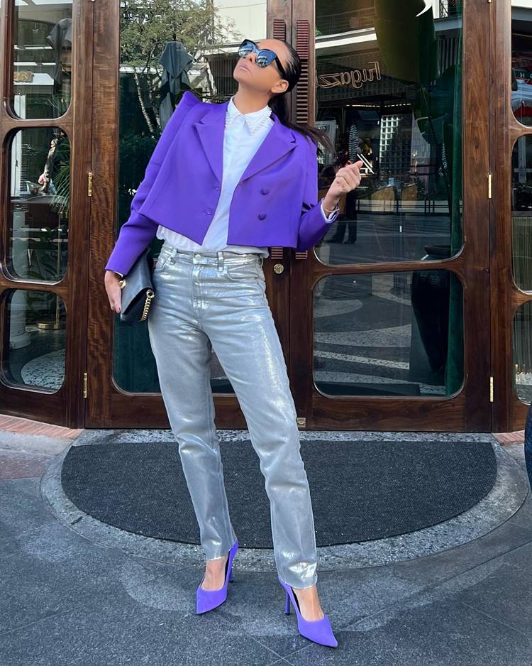 Mulher alta usando camisa branca, blazer lilás curto, calça jeans metalizada, scarpan roxo e bolsa preta, em frente a uma porta de um local público.