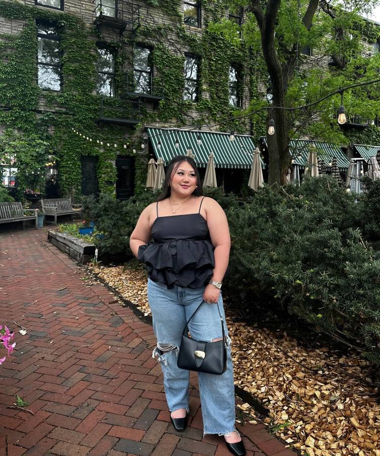 Mulher sorridente com blusa preta de alças, calça jeans rasgada e **sapato preto**, posando em uma calçada de tijolos. Ao fundo, um prédio coberto por hera, árvores e toldos listrados. Ela segura uma bolsa preta.