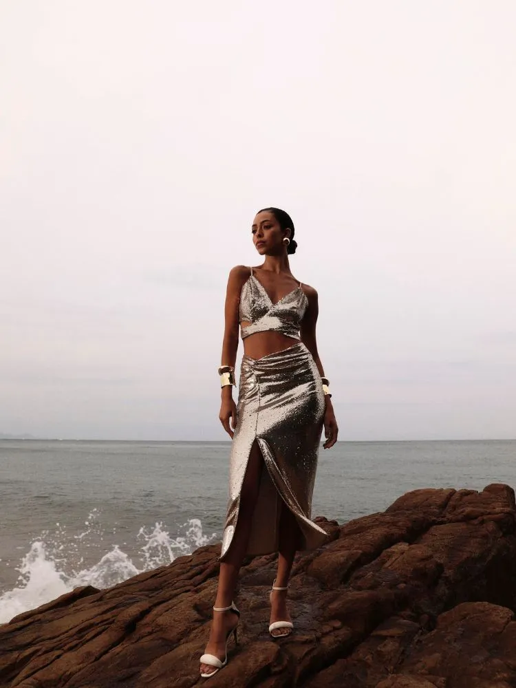 Uma modelo negra possa em uma rocha com o mar ao fundo, ela usa um conjunto de saia de top em tons de cinza e preto da coleção dias dourados da DIMY