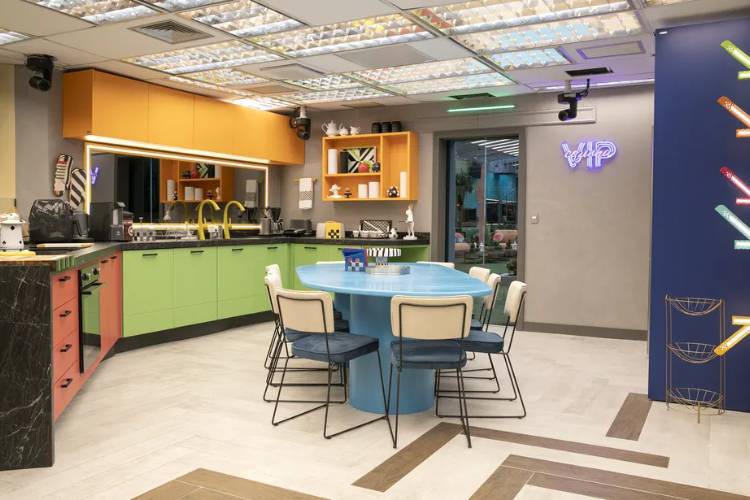 Foto da cozinha do vip do BBB, com mesa azul, cadeiras brancas e azul, armários coloridos e parede cinza. 
