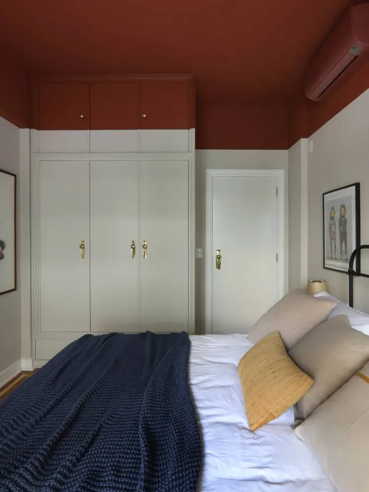 Esta imagem mostra um quarto com um teto colorido em tom terracota, que contrasta com as paredes claras. Uma cama grande com roupa de cama branca e um cobertor azul-marinho domina a cena. Armários brancos com puxadores dourados se estendem até o teto, e uma porta branca é visível ao lado.

