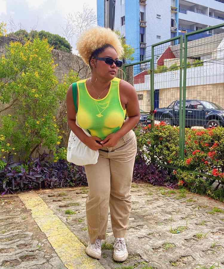 Imagem com fundo de área externa de casa com vista para rua. No centro, mulher usando regata verde e amarela + calça bege + tênis + bolsa branca + óculos de sol + brincos de argola