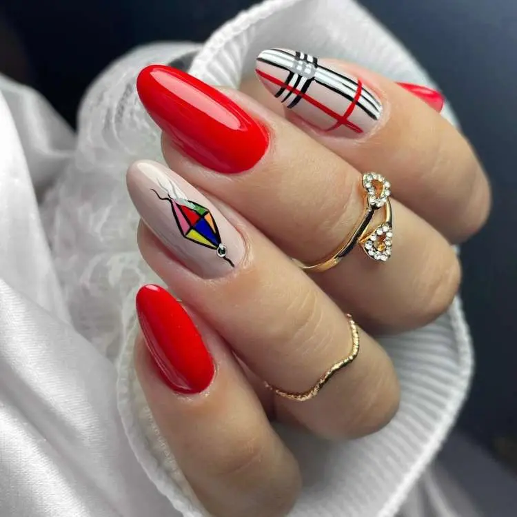 Mão com unhas longas em formato amendoado. Três unhas são vermelhas vibrantes. Duas unhas apresentam padrões de xadrez e uma tem o desenho de uma pipa colorida. Anéis dourados decoram os dedos.