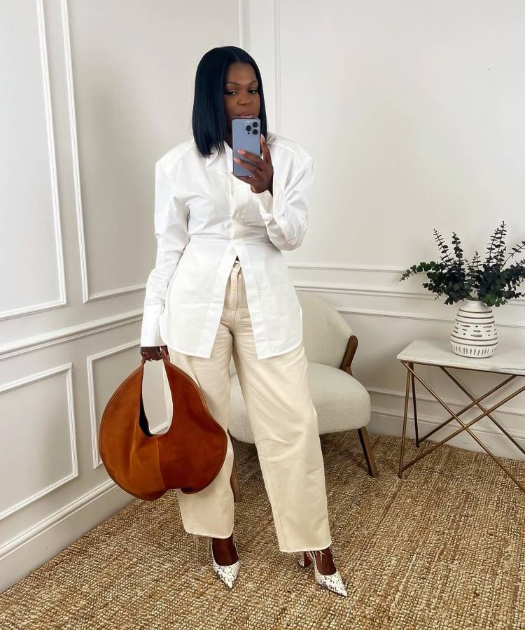 Mulher em ambiente elegante, veste camisa branca oversized, calça bege e sapatos de salto com estampa de cobra. Segura bolsa grande marrom e celular. Ao fundo, cadeira clara, mesa lateral dourada com vaso decorativo e parede branca detalhada.





