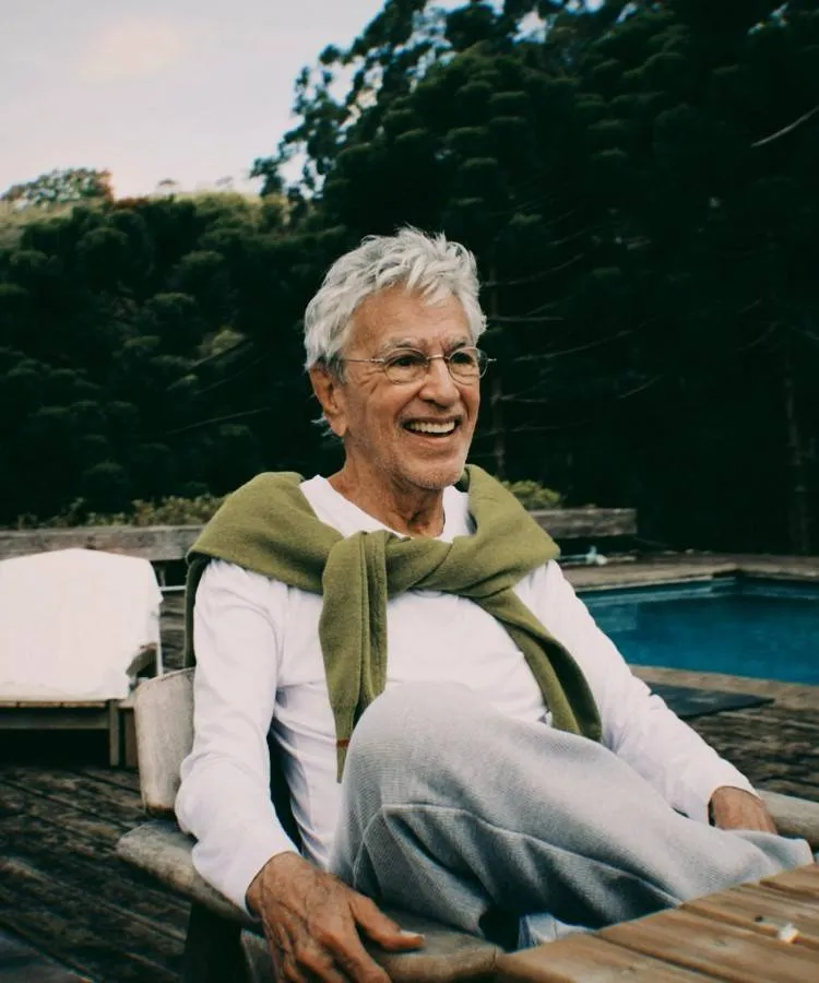 Caetano Veloso entado próximo a uma piscina, veste camisa branca, calças cinza claras e suéter verde sobre os ombros. Ele está relaxado em uma cadeira de madeira, cercado por um ambiente sereno com vegetação e deck rústico.