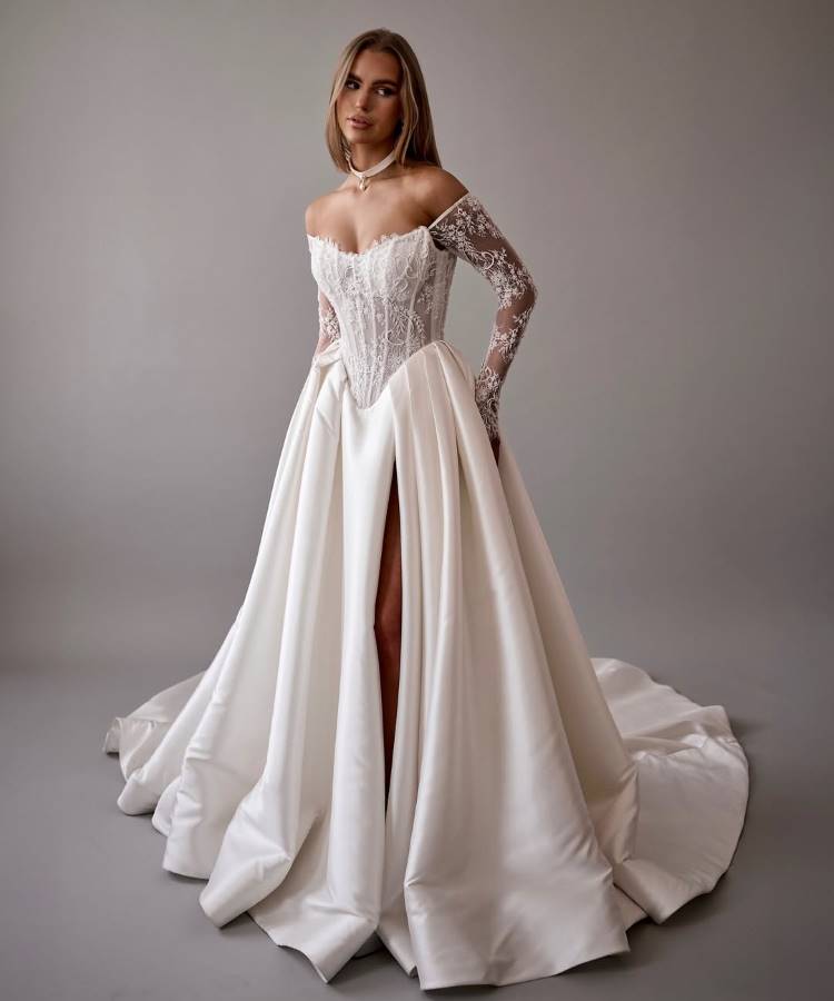 Noiva em vestido branco sofisticado com corpete rendado, decote ombro a ombro, mangas longas de renda e saia volumosa com fenda alta. O design elegante combina delicadeza e modernidade, destacando uma silhueta clássica em um fundo neutro.





