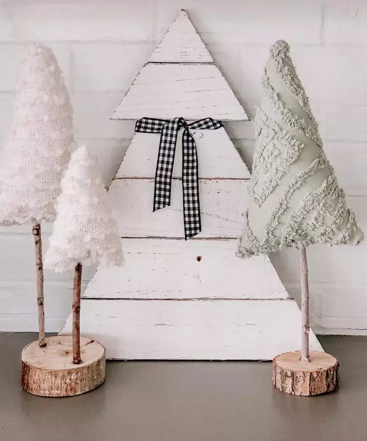 Três árvores decorativas estão sobre uma superfície. Duas são feitas de tecido, uma branca e outra verde, com texturas diferentes, fixadas em troncos de madeira. Ao fundo, uma árvore de madeira branca com laço xadrez preto e branco, encostada em uma parede clara.






