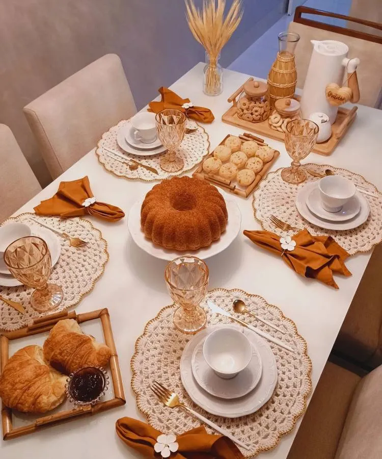 A imagem apresenta uma mesa posta para café da manhã, com sousplats de crochê bege e pratos brancos. Os talheres dourados estão acompanhados de guardanapos laranja com detalhes florais. No centro, um bolo de fôrma redonda está disposto em um prato branco. Ao redor, há croissants com geleia, biscoitos e pães de queijo em bandejas de madeira. Copos de vidro com detalhes dourados e uma garrafa térmica branca completam a cena, criando um ambiente acolhedor e harmonioso.


