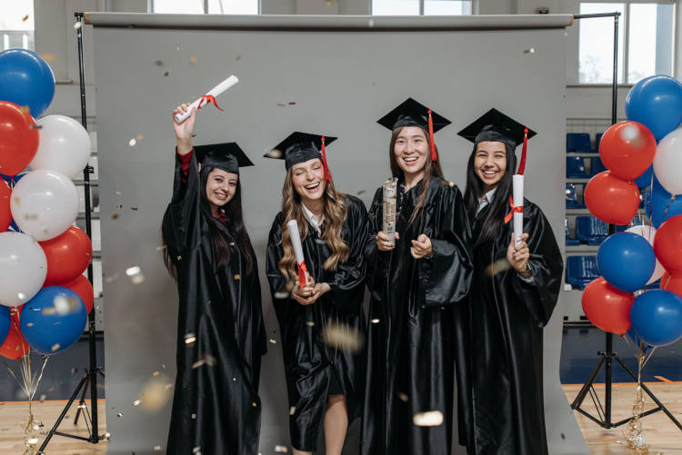Foto de 4 meninas de beca, chapéu e diploma em studio, tirando foto ao lado de bexigas vermelhas, brancas e azuis