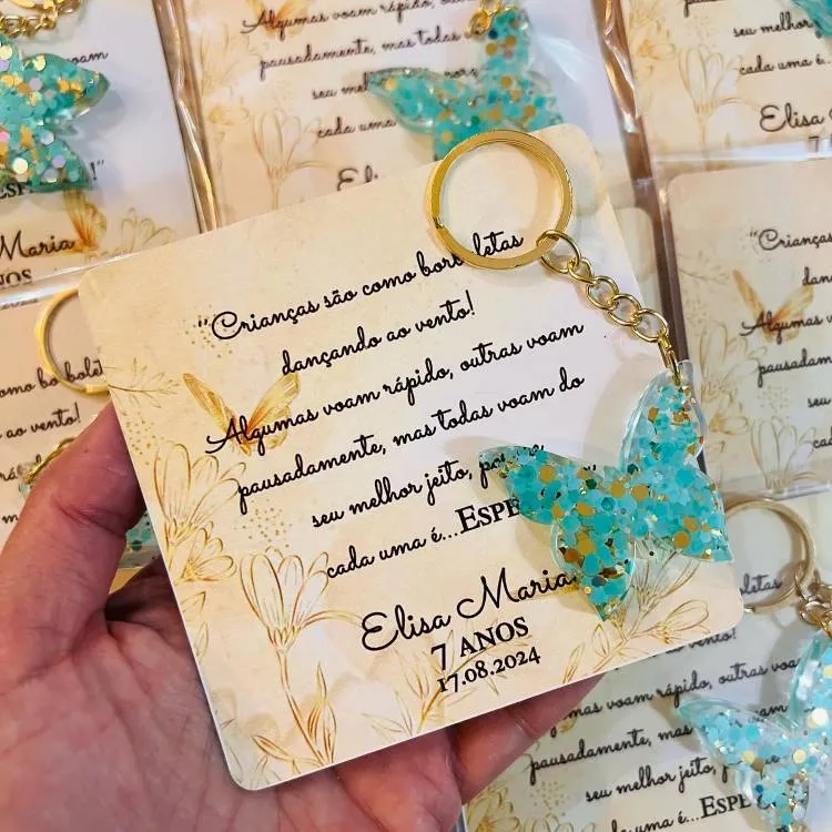 Uma lembrancinha de aniversário que consiste em um cartão com uma citação sobre borboletas e um chaveiro de borboleta de acrílico azul com glitter. O cartão é personalizado com o nome "Elisa Maria".

