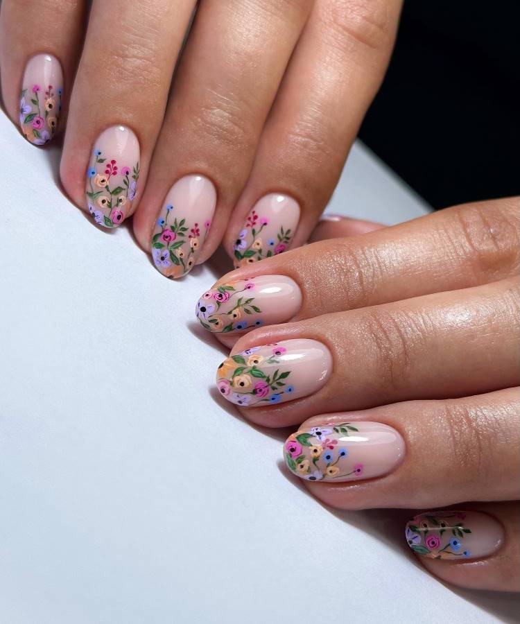 Unhas de gel esmaltadas em nude rosado com acabamento brilhante, decoradas com delicadas flores em tons vibrantes de rosa, laranja, azul e folhas verdes. O design elegante e primaveril combina suavidade e detalhes artísticos, destacando-se pelo visual sofisticado.
