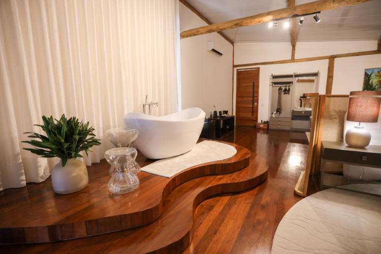 Banheira branca moderna em um quarto rústico e aconchegante. A banheira fica em um tablado de madeira curvilíneo, com teto de vigas expostas e uma grande janela com cortina clara.







