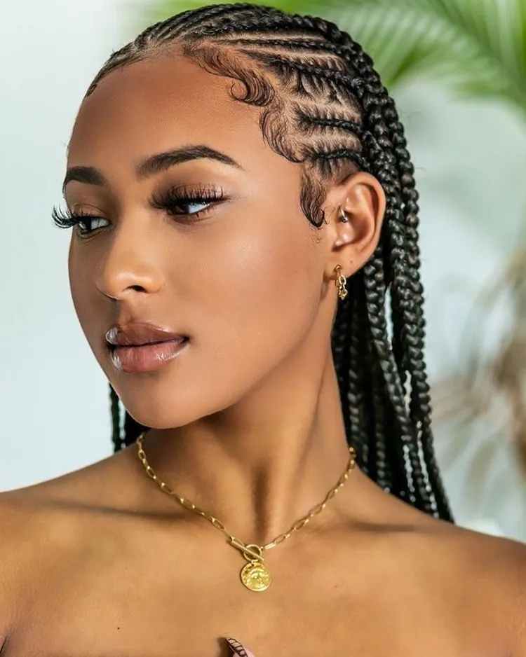 Mulher com trança nagô até metade da cabeça, cabelo todo trançado; Ela tem maquiagem leve e usa brinco e colar dourados