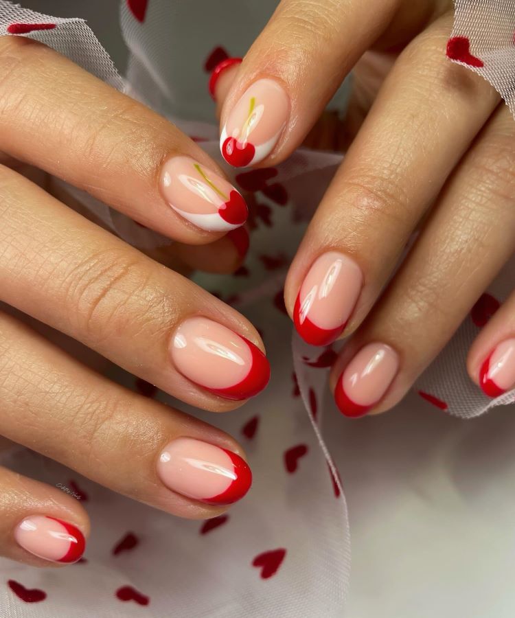 Mãos com unhas decoradas em base nude, com pontas francesas em vermelho vibrante e dois desenhos de cerejas com detalhes em branco e verde. Fundo com tecido branco translúcido decorado com pequenos corações vermelhos, destacando o visual romântico.