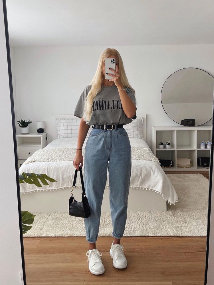 Mulher de pele clara e cabelo loiro usando mom jeans, cinto e camiseta cinza