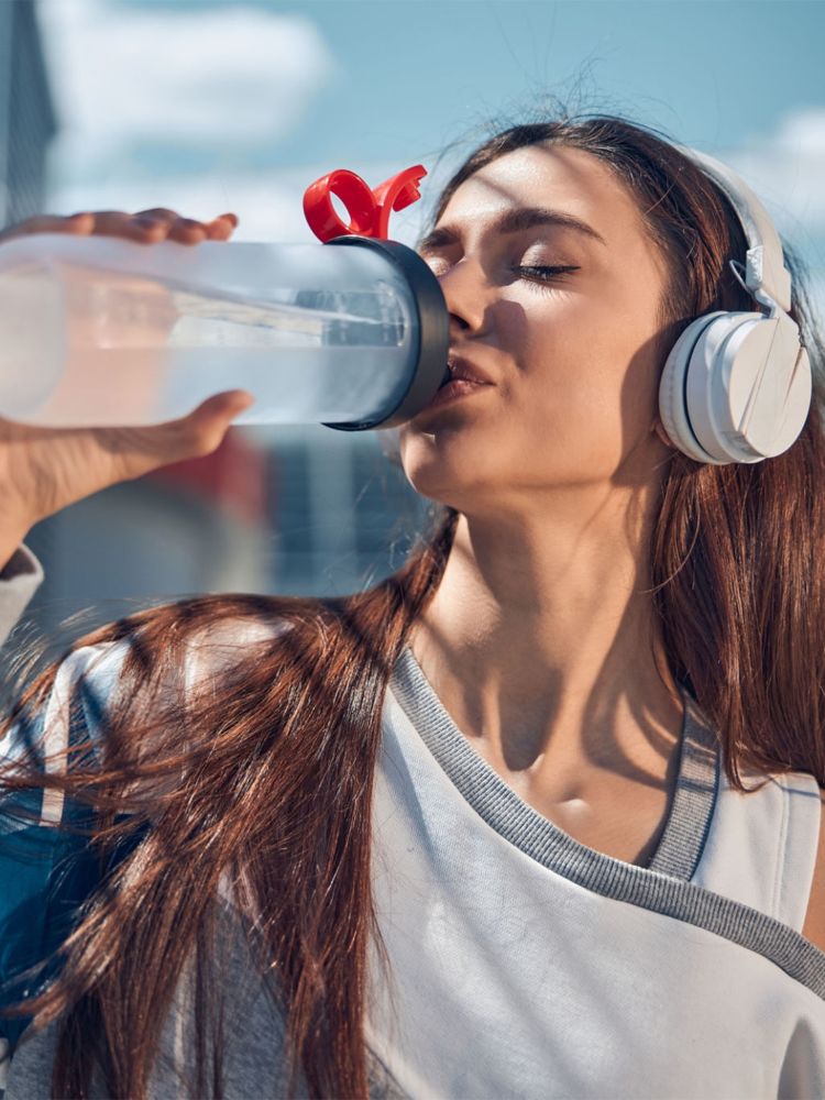 Uma mulher com fones de ouvido brancos, de cabelos castanhos longos, está bebendo água de uma garrafa transparente. Ela tem os olhos fechados e o sol incide sobre seu rosto.