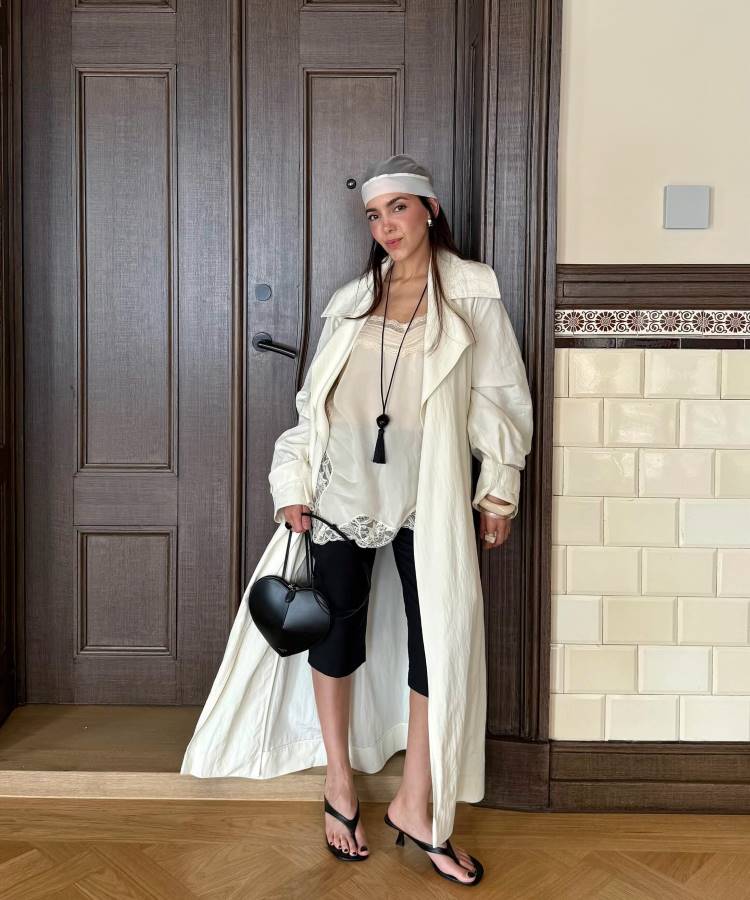 Uma mulher com um turbante cinza e branco posa segurando uma bolsa em forma de coração. Ela veste uma blusa de seda e renda, calça capri preta, um longo sobretudo off-white e chinelos de salto preto.