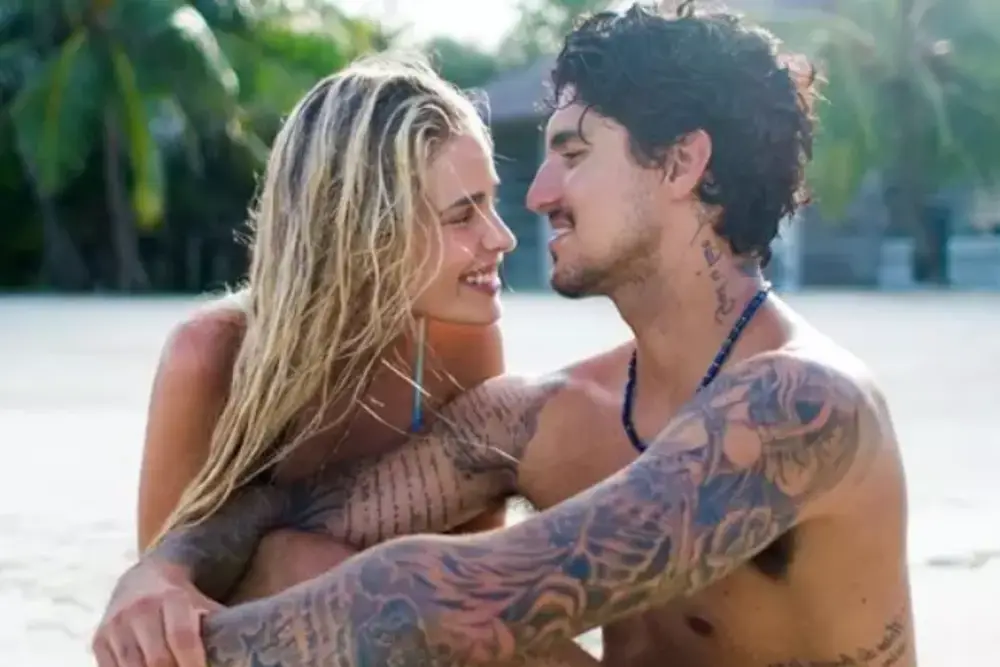 foto de Yasmin Brunet e Gabriel Medina. O casal está sentado na areia de uma praia, os dois estão sorrindo e se olhando nos olhos