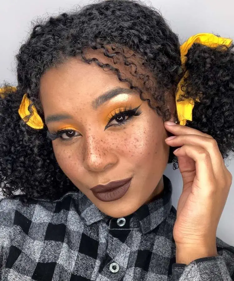 ma mulher com maquiagem de Festa Junina que se destaca pelos tons quentes de amarelo e laranja nos olhos e sardas falsas. Seus lábios têm um batom marrom escuro e o cabelo cacheado está preso em duas marias-chiquinhas com laços amarelos.