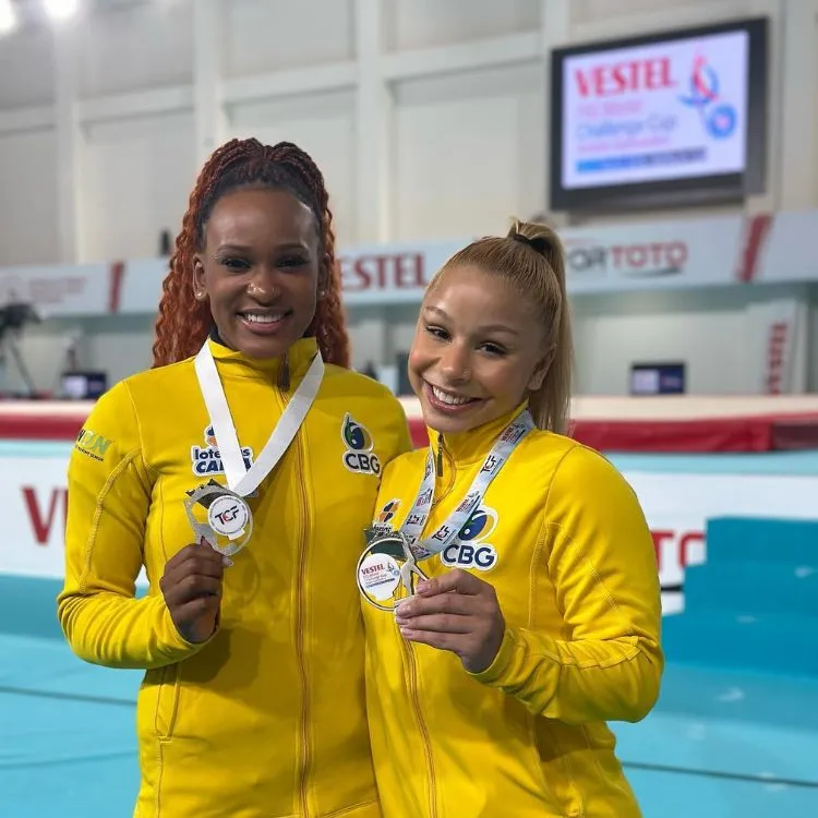 Foto de Rebeca Andrade e Flávia, ambas usando casaco amarelo e segurando medalha
