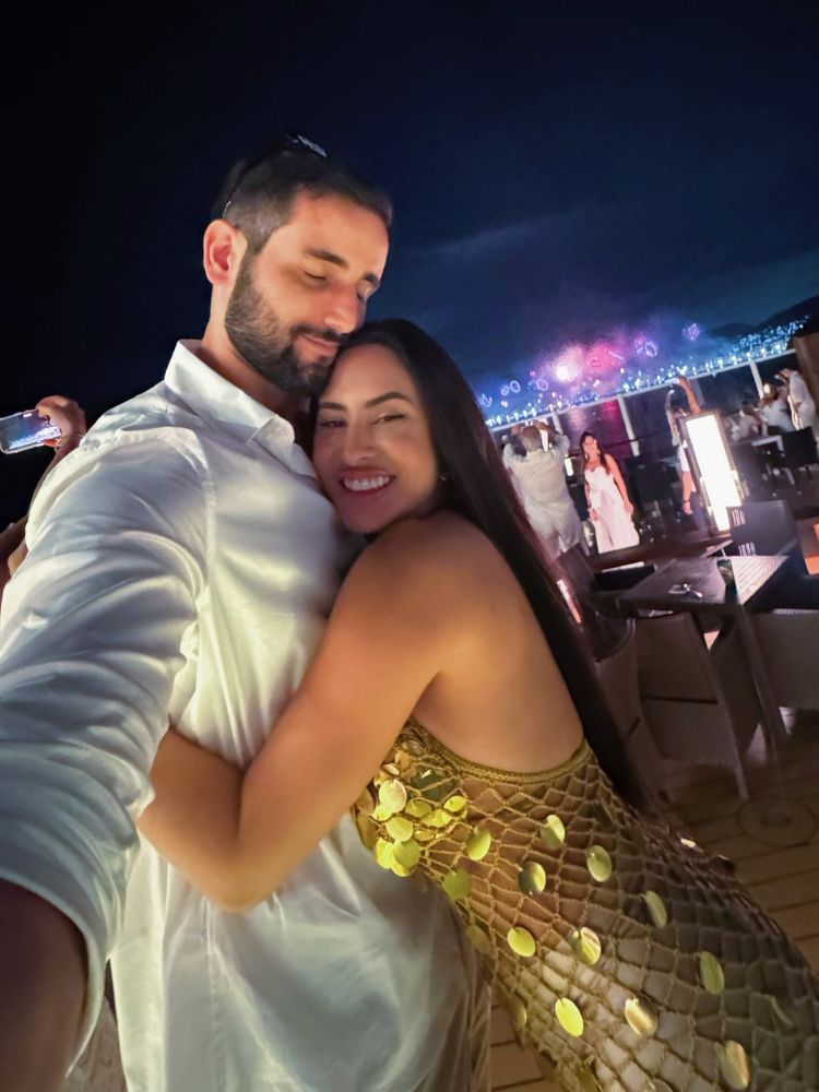 Matteus de camisa branca abraçado com Isabelle de vestido dourado e praia de Copacabana com fogos de artifício ao fundo