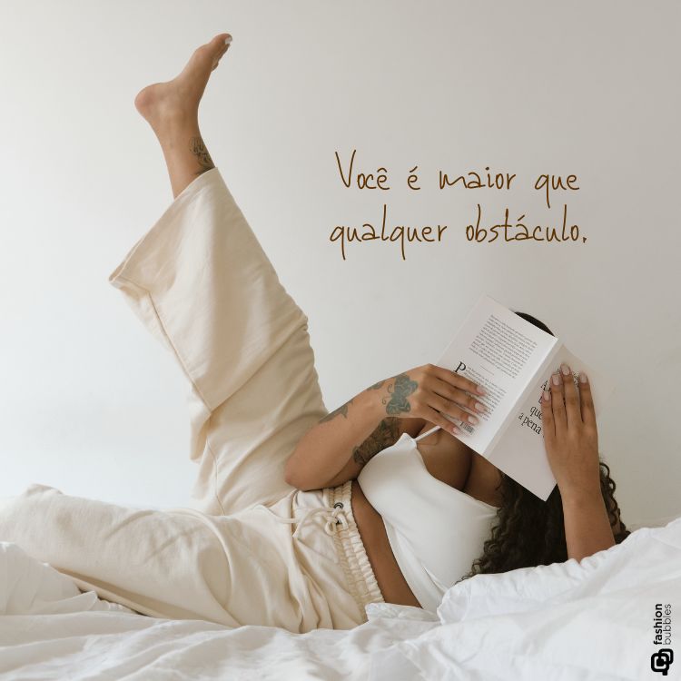 Mulher deitada em uma cama com roupa bege confortável, segurando um livro que cobre parcialmente seu rosto. Ao lado, a frase: "Você é maior que qualquer obstáculo". Imagem inspiradora ideal para divulgar frases de amor-próprio e motivação.