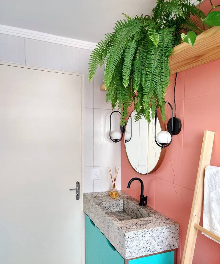 Banheiro decorado com estilo moderno, gabinete azul, pia de granito, parede rosa e iluminação elegante. Uma samambaia exuberante pendurada em prateleira de madeira dá um toque natural ao ambiente, complementado por um espelho oval e toalheiro de madeira.
