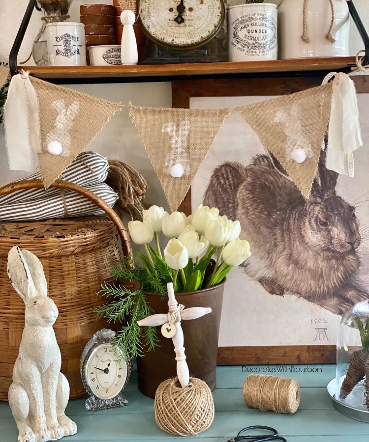 Decoração rústica de Páscoa com guirlanda de juta estampada com coelhos e pompons, flores brancas em vaso metálico, figuras de coelho, cesta de vime, relógio vintage, barbantes e fundo com imagem de lebre; detalhes equilibram simplicidade e charme.