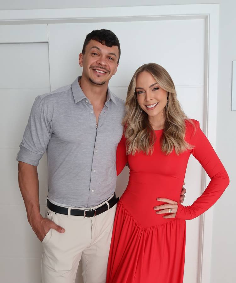 A imagem mostra Tata Estaniecki e seu marido, Júlio Cocielo, lado a lado. Tata veste um vestido longo vermelho vibrante e Cocielo usa uma camisa cinza clara com calças creme. Ambos sorriem para a câmera, com um fundo branco simples.
